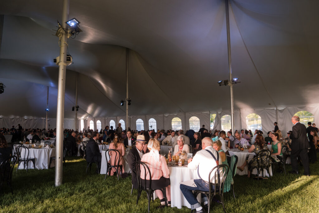Event guests gathered on a lawn with banquet seating for a dinner reception, under a pole tent.