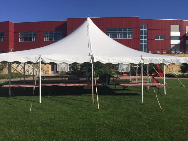 Pole tent with picnic tables underneath, on a lawn in front of a large brick building.