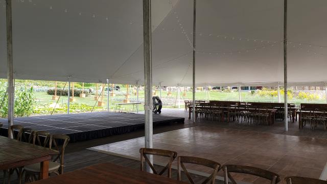 Rectangular tables, wooden dance floor, and a stage rental under a pole tent with string lights.