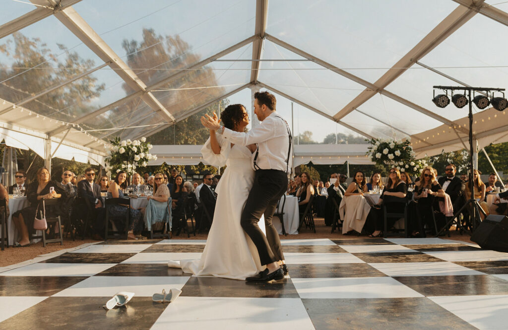 Bride and Groom slow dancing on a black and white dance floor rental, while their guests watch at tables under a frame tent.