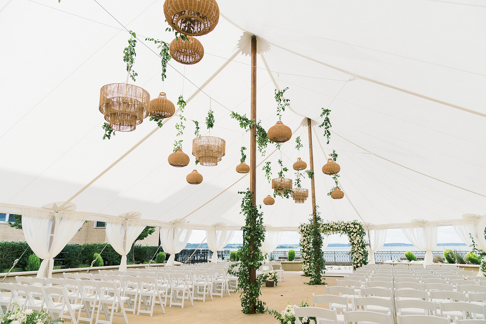 Chair rentals set up for a wedding ceremony under a sailcloth tent, decorated with hanging light fixtures and greenery.
