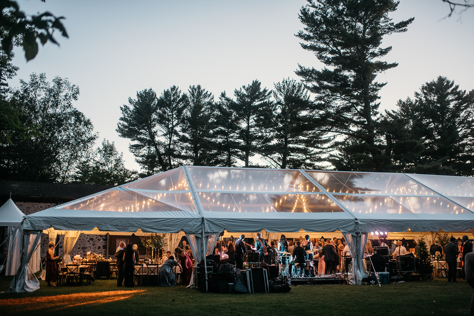 A group of event guests watching a band play under a frame tent with string lights at evening time.
