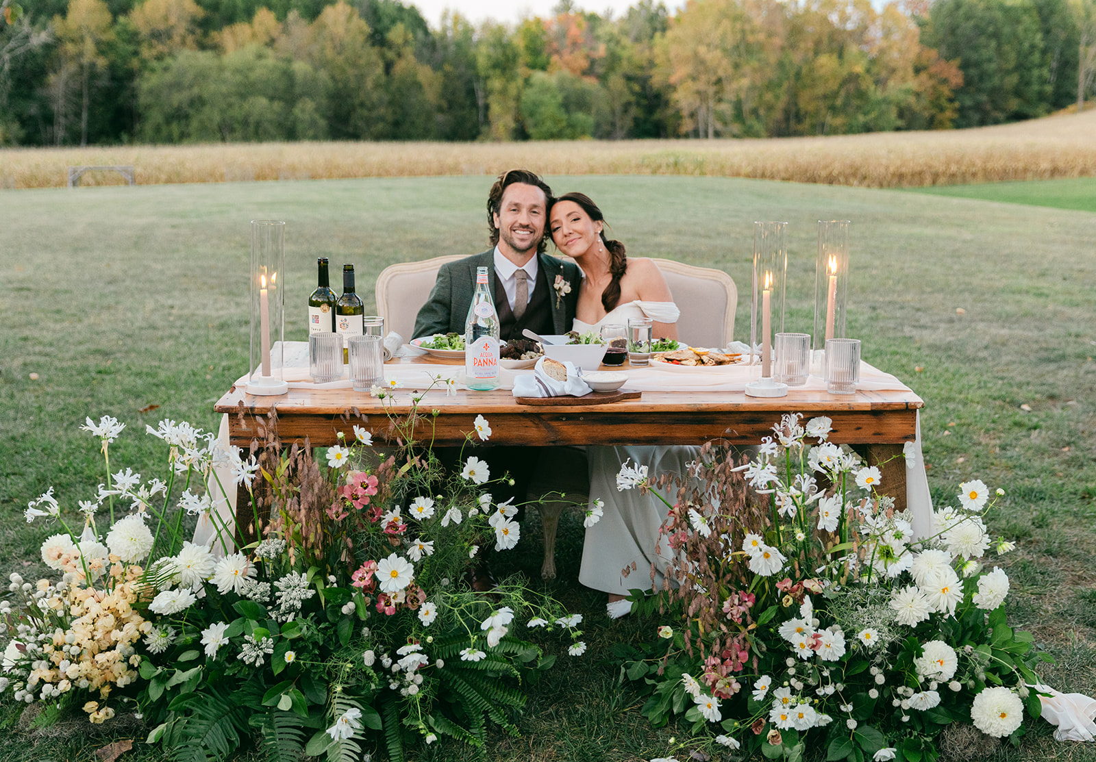 Bride and Groom enjoying their wedding meal at a table with bottles of wine and candles, at an outdoor venue.